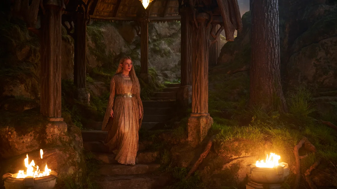 Uma elfa de vestido dourado caminhando entre árvores e tochas, numa floresta élfica, em imagem oficial da 3ª temporada de Os Anéis de Poder