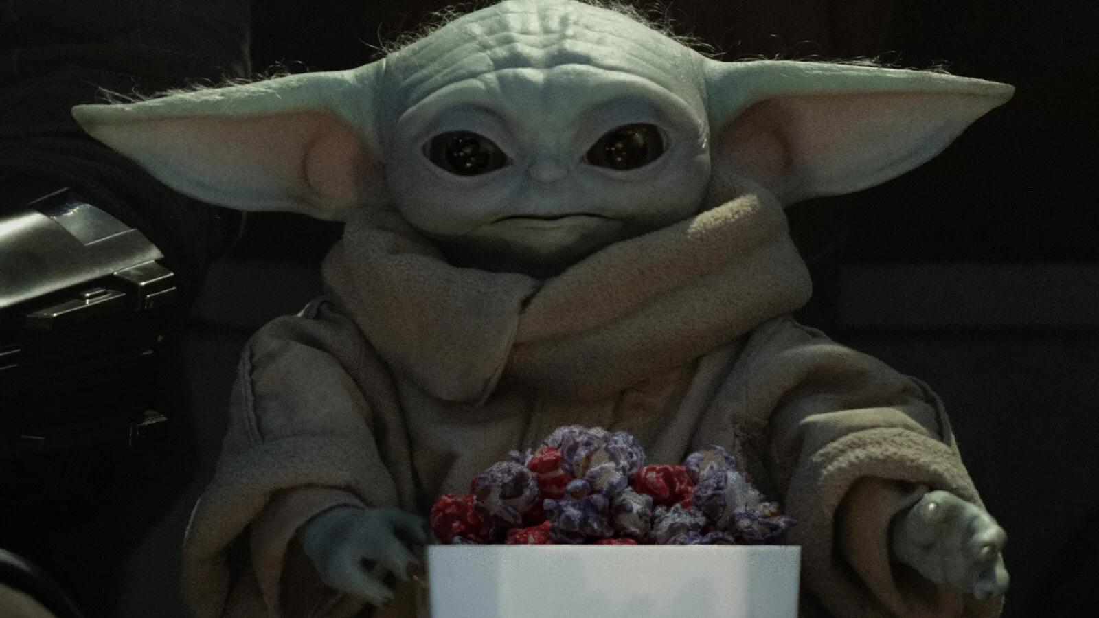 Grogu, o pequeno da espécie do Yoda, segurando um balde de pipoca, em imagem oficial do guia de filmes e séries do StarWars.com